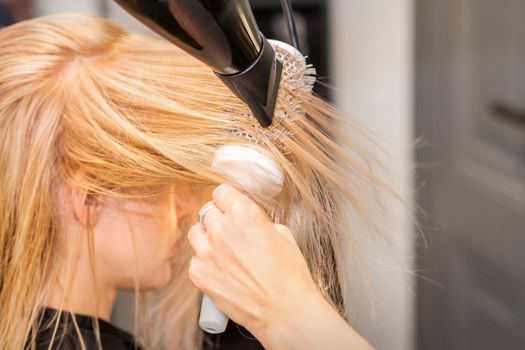 Drying straight blond hair with black hairdryer and white round brush in hairdresser salon, close up