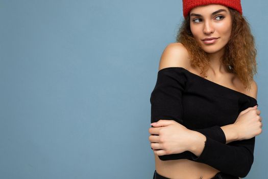 Photo of pretty positive sexy young brunet curly woman isolated over blue background wall wearing black crop top and stylish red hat looking to the side. Copy space