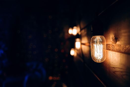 Antique incandescent Edison type bulb on wooden texture wall. Blurred warm garland light on background. Christmas night party mood. Copy space for congratulations. Festival interior decorating.