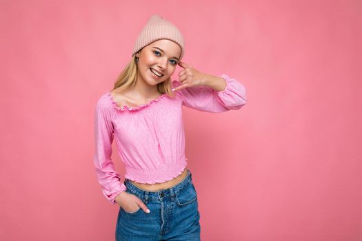 Photo of beautiful happy smiling young blonde woman isolated over pink background wall wearing trendy pink hat and pink blouse looking at camera and immitating communication via mobile phone.