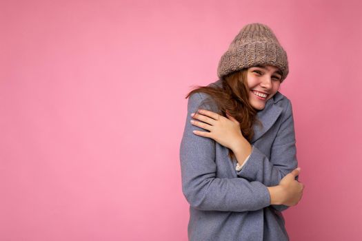 Photo of beautiful positive funny joyful smiling young brunette woman isolated over pink background wall wearing grey autumn coat and grey warm hat looking at camera and feeling cold. Free space, copy space