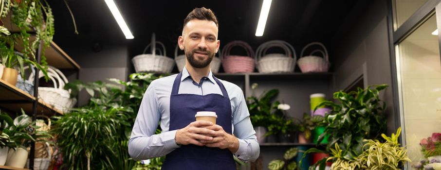 a successful man florist with a cup of coffee in his hands stands in a flower shop looking at the camera with a smile on his face.
