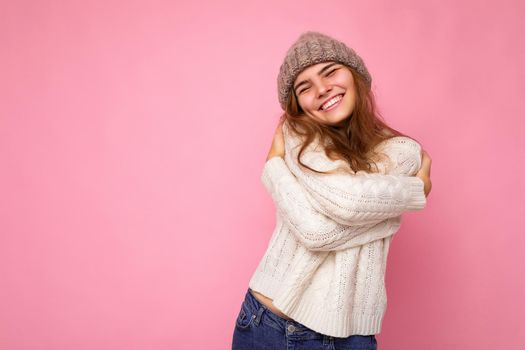 Shot of beautiful happy positive smiling young brunette woman isolated over pink background wall wearing knitted white sweater and grey warm hat and having fun. Free space