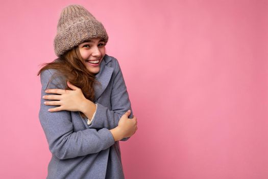 Shot of fascinating happy smiling young brunette woman isolated over pink background wall wearing grey autumn coat and grey warm hat looking at camera and feeling cold. Empty space, copy space