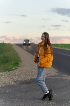Cool modern teen girl poses on a lonely road while the sky background looks blue with clouds at sunset.
