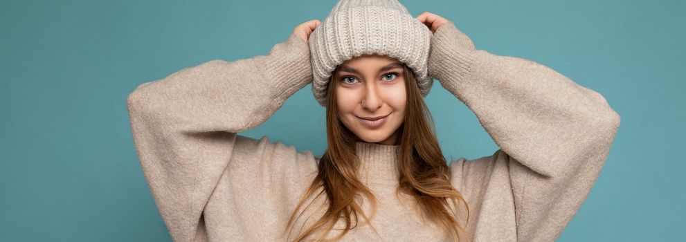 Panoramic photo of attractive pretty beautiful positive happy smiling young dark blond woman isolated over blue background with empty space wearing beige warm sweater and knitted beige hat looking at camera.