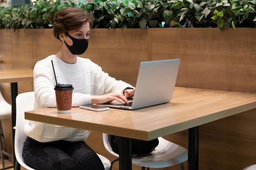 freelancer girl in a shopping center at the food court works behind a laptop in a mask from covid.
