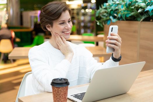 Young beautiful pretty positive brunette woman wearing white sweatshirt sitting at a shopping center at a table and working at a computer laptop, using mobile phone. Freelance and business concept.