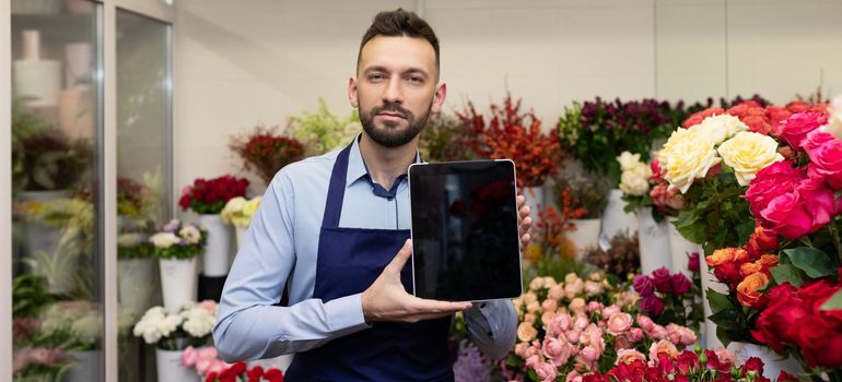 a man florist in a flower shop stands among the bouquets with a tablet in his hands with the screen forward.