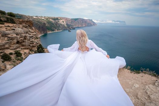 Blonde with long hair on a sunny seashore in a white flowing dress, rear view, silk fabric waving in the wind. Against the backdrop of the blue sky and mountains on the seashore