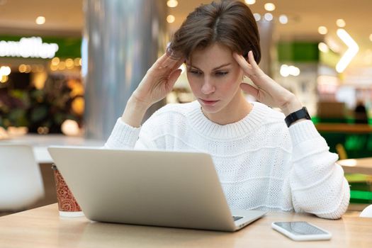 Young beautiful thinking brunette woman wearing white sweatshirt sitting at a shopping center at a table and working at a computer laptop. Freelance and business concept.