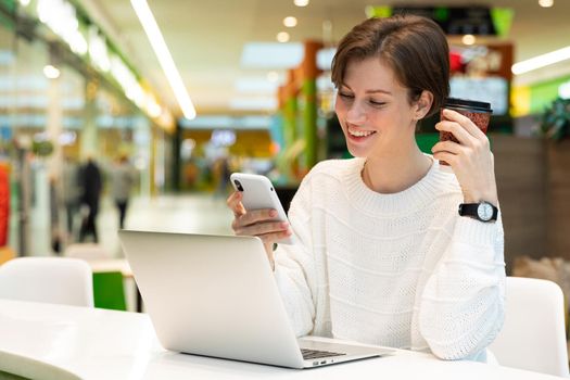 Young beautiful woman sitting at a shopping center at a table and working at a computer laptop, using mobile phone. Freelance and business concept.