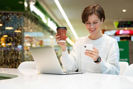 Young beautiful woman sitting at a shopping center at a table and working at a computer laptop, using mobile phone, drinking coffee. Freelance and business concept.