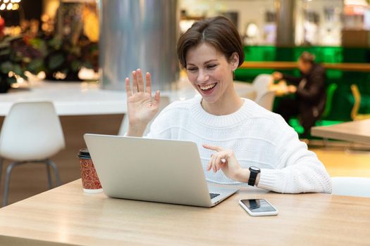 Young beautiful smiling happy brunette woman wearing white sweatshirt sitting at a shopping center at a table and working at a computer laptop. Freelance and business concept.