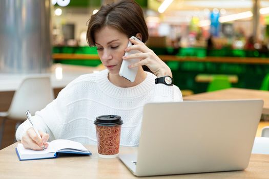 Young beautiful pretty brunette woman wearing white sweatshirt sitting at a shopping center at a table and working at a computer laptop, using mobile phone, writing information at notebook. Freelance and business concept.