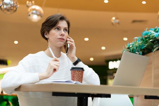 Young attractive brunette woman sitting at a shopping center at a table and working at a computer laptop, using mobile phone, writing information at notebook. Freelance and business concept.