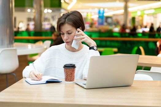 Young beautiful pretty brunette woman wearing white sweatshirt sitting at a shopping center at a table and working at a computer laptop, using mobile phone, writing information at notebook. Freelance and business concept.