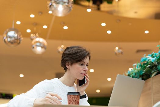 Young attractive brunette woman sitting at a shopping center at a table and working at a computer laptop, using mobile phone. Freelance and business concept.