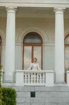 a beautiful mature woman in a long white dress stands on the balcony and looks into the distance.