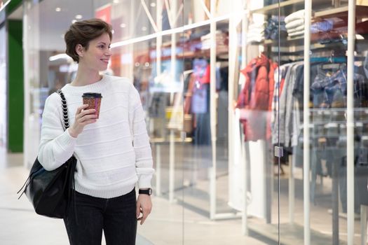 a woman walks through the mall with a glass of coffee in her hands, looks at the windows.