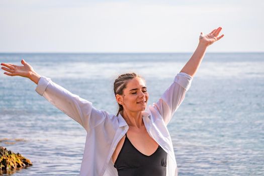 The girl stands on the shore and looks at the sea. Her hands are raised up. She wears a white shirt and her hair is in a braid