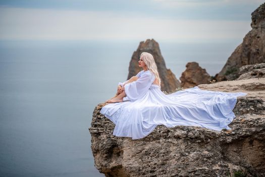 The blonde sits on a rock above the sea. With long hair on a sunny seashore in a white dress, rear view, silk fabric flutters in the wind. Against the backdrop of blue skies and mountains