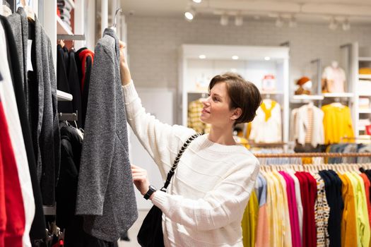 Photo of young attractive brunette woman with a short haircut in a white sweater chooses stylish and casual clothes in a store in a shopping mall. Shop concept.