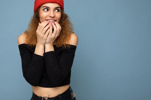 Shot of young emotional positive amazed surprised nice winsome brunet woman wavy-haired with sincere emotions wearing black crop top and red hat isolated on blue background with free space.