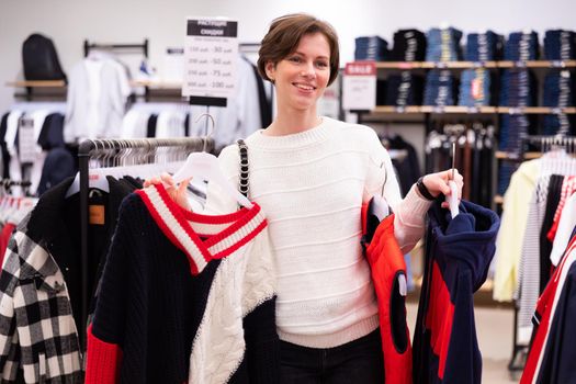 Photo of adult positive smiling attractive brunette woman with a short haircut in a white sweater chooses stylish and casual clothes in a store in a shopping centre. Shop interior.