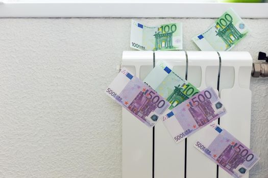 Paper banknotes of 100 and 500 euros on a small white radiator for heating the house. The concept of expensive heating costs and the energy crisis in Europe.