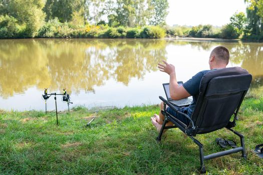 A man sits in a chair near the autumn lake and works on a laptop freelance camping. Robot out of the office, phoned with colleagues