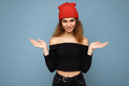 Photo of young positive happy attractive brunette curly woman with sincere emotions wearing black crop top and red hat isolated on blue background with empty space and having doubts.