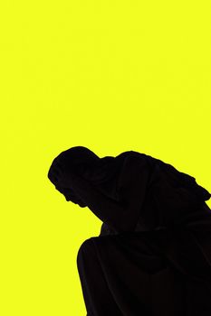 Statue shadow silhouette of an angel holding head with hand, against a strong yellow background.