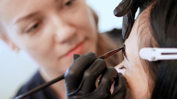 close up. young woman gets eyebrow correction procedure. kosmetolog- makeup artist in black rubber gloves applies paint with brush on eyebrows in beauty saloon. Professional care for face. High quality photo