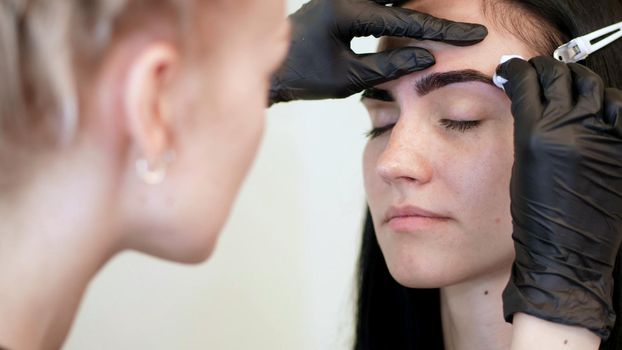 beauty saloon. cosmetologist, in black rubber gloves, gently wipes his eyebrows with a cotton disc, cotton pad, removes the remnants of paint after dyeing his eyebrows. High quality photo