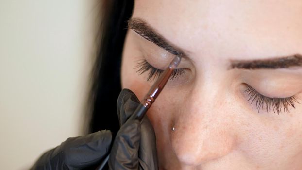 close up. young woman gets eyebrow correction procedure. kosmetolog- makeup artist applies paint with brush on eyebrows in beauty saloon. Professional care for face. High quality photo