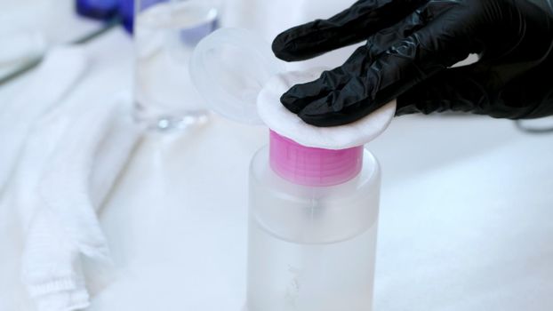 close-up, the hands of the beautician in black rubber gloves hold the cotton pad and moisten it in a special transparent liquid from the bottle with a dispenser. High quality photo