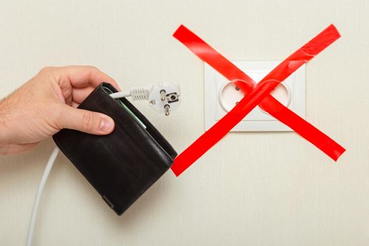 A mans hand holds a black wallet with a white cord hanging from it with a plug near an electrical outlet, tied with a red ribbon crosswise. The concept of the energy crisis in Europe and the increase in the cost of energy carriers