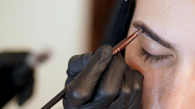 close up. young woman gets eyebrow correction procedure. kosmetolog- makeup artist applies paint with brush on eyebrows in beauty saloon. Professional care for face. High quality photo