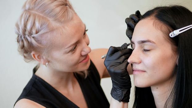 beauty saloon. the cosmetician in black rubber gloves hold tweezers and pull out eyebrows. Master corrects the shape of the eyebrows. near a large mirror with illumination. High quality photo
