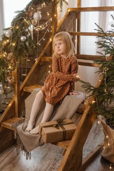 A little blonde girl is sitting on a wooden staircase in a Scandinavian interior decorated in a New Year's style. A child holds a Christmas gift in a craft package.