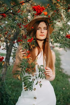 A beautiful blonde girl in a hat with long hair, on a walk in the autumn season. Portrait of a woman at a rowan tree.