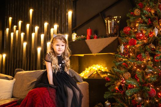 A girl in a festive outfit on the sofa with her dog next to a Christmas tree decorated with garlands, balloons and Christmas toys. The concept of winter holidays is Christmas and New Year holidays. Magical festive atmosphere.