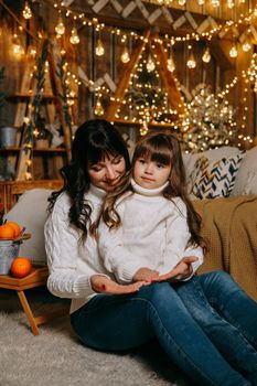 A little girl with her mother in a cozy home environment on the sofa next to the Christmas tree. The theme of New Year holidays and festive interior with garlands and light bulbs