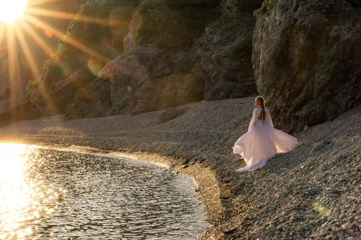 A mysterious female silhouette with long braids stands on the sea beach with mountain views, Sunset rays shine on a woman. Throws up a long white dress, a divine sunset