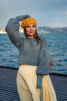 Outdoors fashion portrait of a beautiful middle aged woman walking on the beach. Marine background. Dressed in a stylish warm blue sweater, yellow skirt and beret