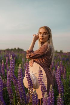 A beautiful woman in a straw hat walks in a field with purple flowers. A walk in nature in the lupin field.