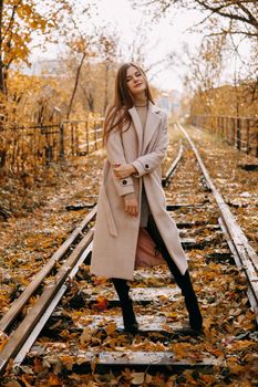 A beautiful long-haired woman walks through the autumn streets. Railway, autumn leaves, a woman in a light coat.