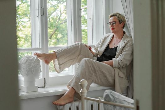 A middle-aged woman in a beige suit and black tank top sits mysteriously and looks out the window on the windowsill. Green trees outside.