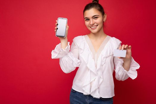 Portrait of positive cheerful fashionable woman in formalwear holding and showing mobile phone with empty screen for mock up and credit card with cut out looking at camera isolated on red background with copy space.